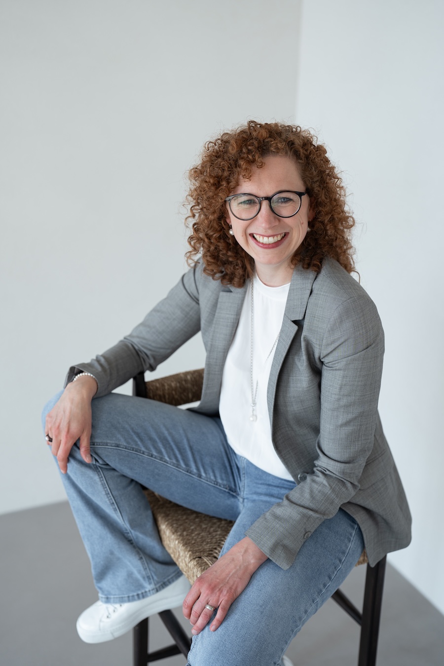 „Sarah Vögelin, Inhaberin von Room and Color, Porträt im grauen Blazer und Jeans auf Stuhl vor hellem Hintergrund, von Room and Color in Hölstein (Basel-Landschaft), Schweiz